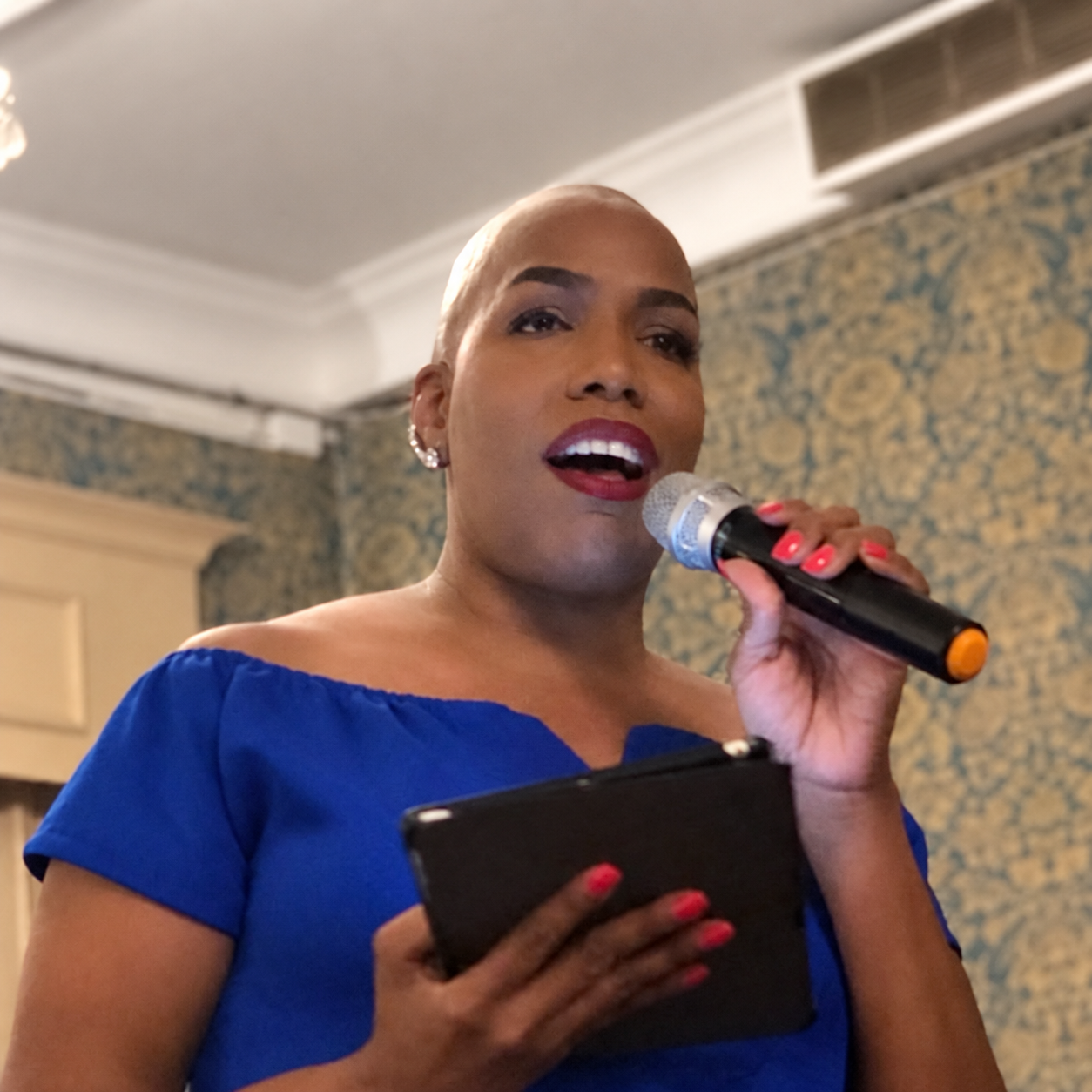 Sue A. Webley in blue, holding a microphone and tablet while speaking